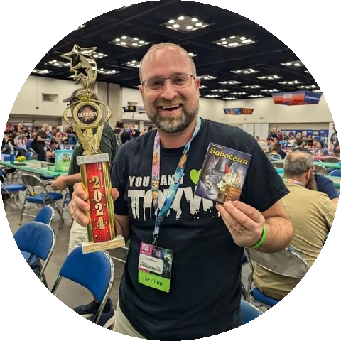 Jason Kogan at Gen Con holding a Saboteur championship trophy and a Saboteur game box